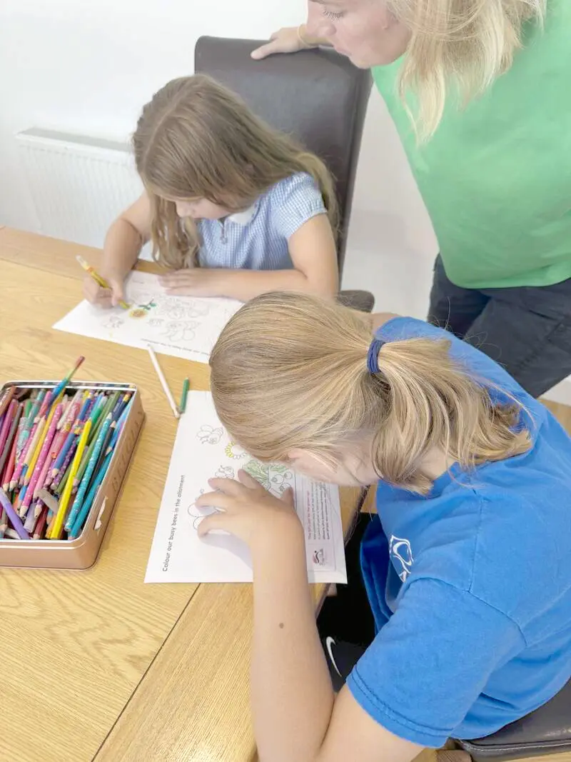Two children colouring in a Bookbag activity sheet at the kitchen table, with a parent looking on